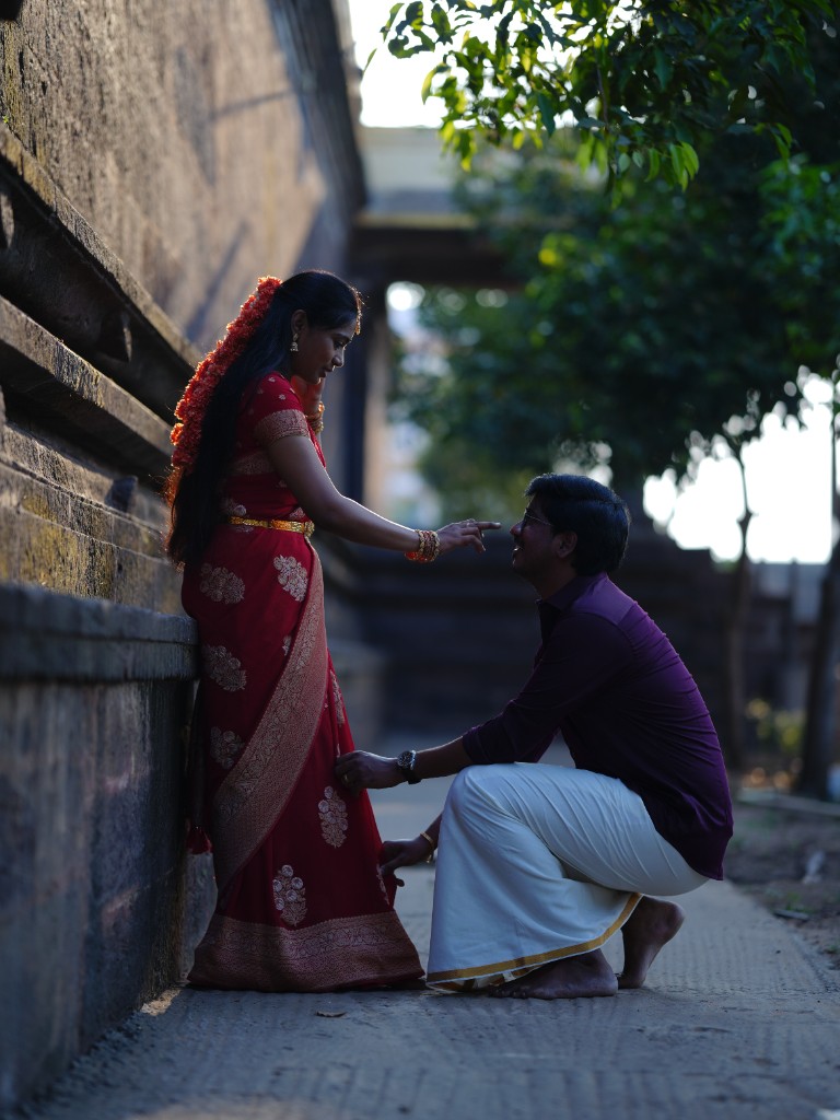 Srinu kneeling and adjusting Sai's saree beside the temple wall